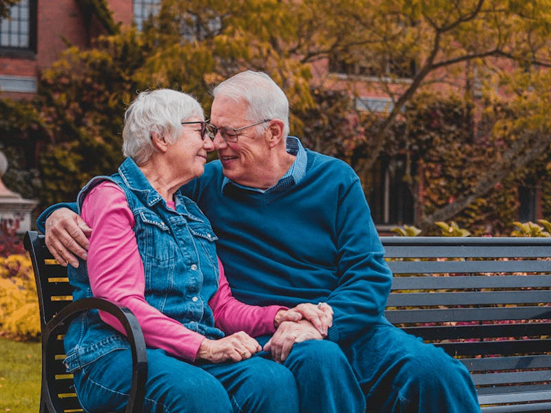 An elderly couple sitting on a park bench in the autumn.